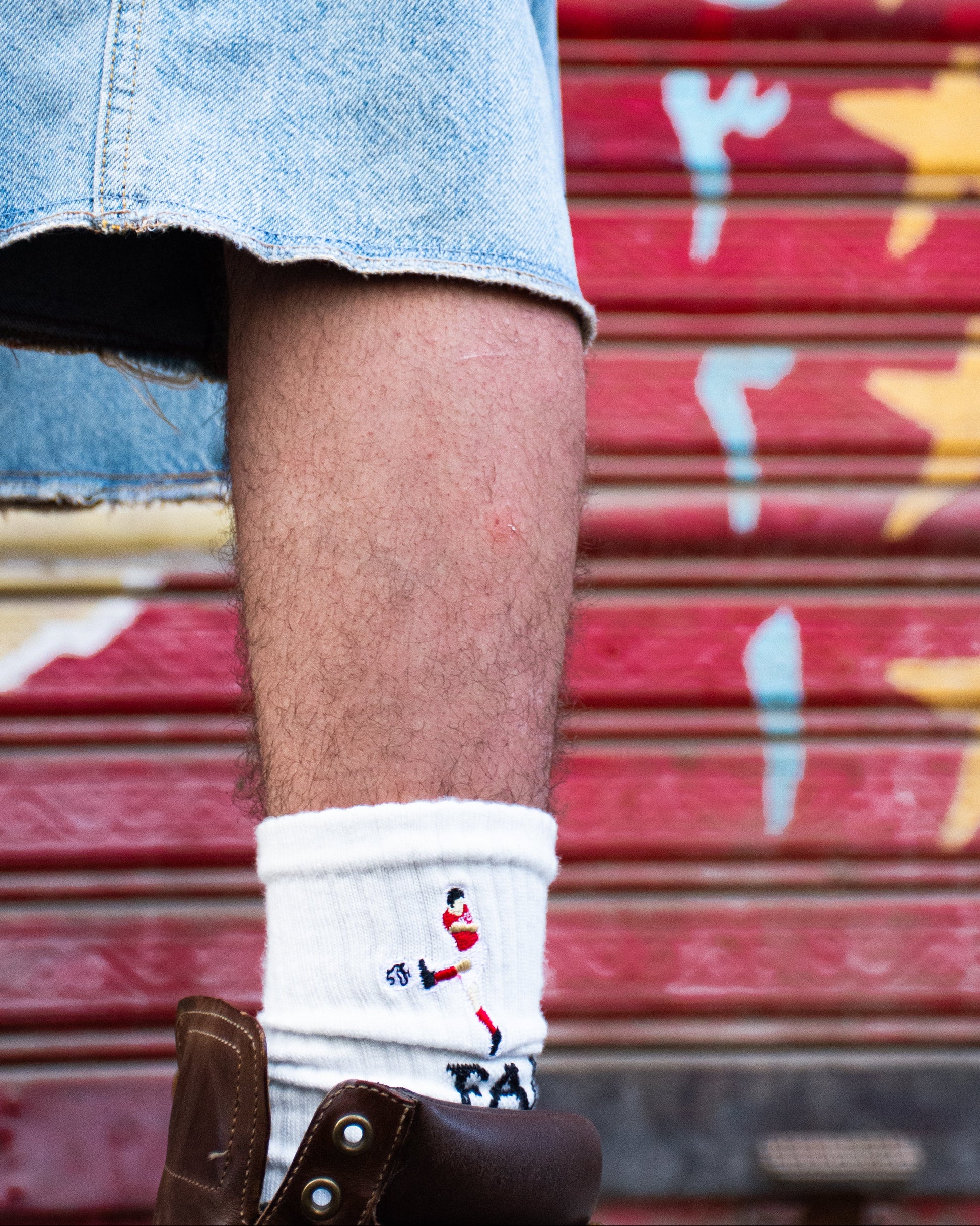 Person wearing denim shorts and a white sock with a design, standing in front of a red shutter with yellow star patterns.